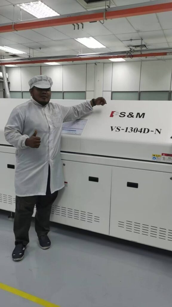 A worker in overalls gives a thumbs-up next to an S&M Corp. VS-1304D-N reflow oven.